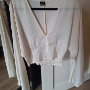 Vit croppad blus från Gina Tricot - Säljer en vit croppad blus från Gina Tricot med v-ringning och långa, vida ärmar. Blusen har smockad nederkant som ger en snygg passform och är tillverkad i ett lätt, flowy material. Perfekt för dig som gillar stilrena och trendiga plagg.