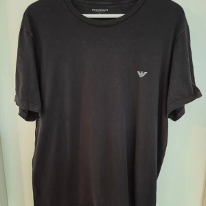 Svart t-shirt från Emporio Armani XL - Svart t-shirt från Emporio Armani med diskret logga på bröstet. Klassisk rund halsringning och korta ärmar. Tillverkad i mjuk bomull som känns skön mot huden. Perfekt för en clean och stilren look.