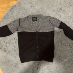 Grå och svart kofta från Skill - Snygg kofta från Skill i tvåfärgad design med gråmelerad ovandel och svarta partier nedtill och på ärmarna. Tröjan har långa ärmar, ribbade muddar och knäppning framtill med svarta knappar. Perfekt för lager-på-lager och chill stil.