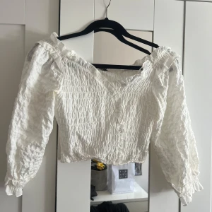 Vit smockad blus med volang - Supersöt vit blus med smockad struktur och volangdetaljer vid axlarna. Blusen har v-ringning och långa ärmar med resår vid ärmslut. Perfekt till jeans eller kjol för en fräsch look.