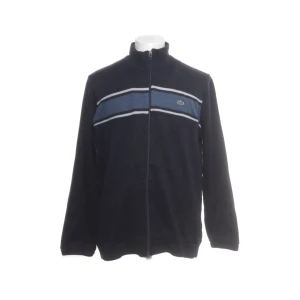 Mörkblå kofta från Lacoste med dragkedja - Lacoste Tröja - Storlek M (Herr)  Klassisk och stilren tröja från Lacoste med dragkedja framtill. Perfekt för dig som vill kombinera sportig design med tidlös elegans och komfort.  Storlek: M (herr)  Färg: Mörkblå med blå/vita ränder över bröstet  Full dragkedja framtill  Hög krage för extra komfort och  stil  Diskret Lacoste-logga på bröstet  Material: Mjuk och bekväm, passar både vardag och fritid