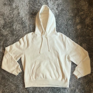 Vit hoodie med röda läppar på ryggen -  Vit hoodie i bomull med huva och snörning. På ryggen finns ett egen gjort stort tryck med röda läppar i ett rektangulärt mönster. Klassisk känguruficka framtill och ribbade muddar vid ärmslut och nederkant. Perfekt för dig som gillar unika detaljer.