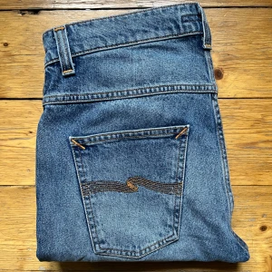 Blå jeans från Nudie Jeans straight fit - Säljer ett par klassiska blå jeans från Nudie Jeans med raka ben och snygga kontrastsömmar. Jeansen har fem fickor och den ikoniska Nudie-bågen på bakfickorna. Tillverkade i slitstark bomullsdenim och passar perfekt till en avslappnad stil. Priset är inte hugget i sten!  W32 L34