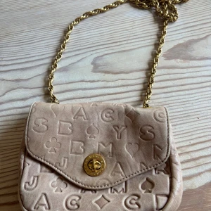 Beige axelväska Marc by Marc Jacobs - Söt beige axelväska i skinn från Marc by Marc Jacobs med präglade bokstäver och symboler. Väskan har guldfärgad kedjerem och ett snyggt vridlås. Insidan är fodrad med tyg i vinrött och röda läppar. Perfekt storlek för det viktigaste.