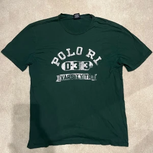 Grön t-shirt från Polo Ralph Lauren - Snygg grön t-shirt från Polo Ralph Lauren med tryck 'Polo RL 033 Varsity Tr.' i vitt på framsidan. Tillverkad i 100% bomull, perfekt för en avslappnad stil. Det står L men sitter som XL. Vid minsta fråga eller fundering hör av er🤝