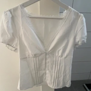 Vit blus med spets och knappar - Söt vit blus med kort puffärm, djup v-ringning och spetskant längs urringningen. Framsidan har dekorativa knappar och fina veckade detaljer.