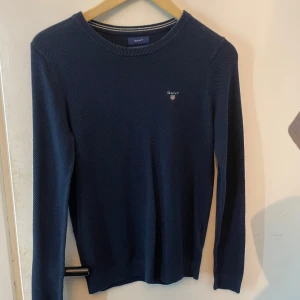 Mörkblå stickad tröja från Gant - Säljer en mörkblå stickad tröja från Gant med rund halsringning och diskret logga broderad på bröstet. Tröjan har använts ett par fåtals gånger. Den har lite fjun på sig vilket syns på bilden, den kommer såklart tvättas och det är inga problem med att be om bild efteråt! 