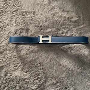H skärp med H-spänne vändbart - Säljer två stilrena H bälten i skinn, ett i marinblått och ett i svart. Båda har det ikoniska H-spännet i silverfärg och vit kontrastsöm längs kanterna. Perfekt accessoar för att lyfta vilken outfit som helst.