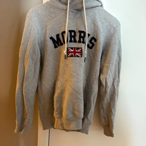 Grå hoodie från Morris med tryck - Snygg grå hoodie från Morris med svart text och brittisk flagga på bröstet. Tröjan har huva med vita snören och en stor magficka. Perfekt för dig som gillar klassisk stil.