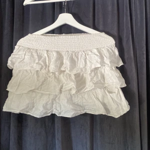 Vit volangkjol från Lindex - Supersöt vit kortkjol från Lindex med flera lager volanger och bred resår i midjan. Kjolen är gjord i mjuk bomull och har en luftig känsla, perfekt för varma dagar. Enkel att matcha med både toppar och sneakers.