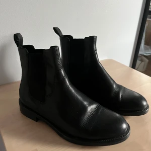 Svarta chelsea boots från Vagabond - Snygga svarta chelsea boots från Vagabond i äkta skinn med elastiska sidor och dragflikar både bak. Klassisk rund tå och platt sula ger en stilren vibe som funkar till allt. Perfekt för dig som gillar minimalistisk och tidlös stil. Använda några gånger men fortfarande i bra skick och inga defekter.