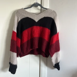 Randig stickad tröja i rött och svart - Snygg stickad tröja med breda ränder i rött, svart, vinrött och beige. Tröjan har en loose passform, ribbad rund hals och långa ärmar med kontrastfärgade muddar. Perfekt för dig som gillar färg och vill ha en chill vibe.
