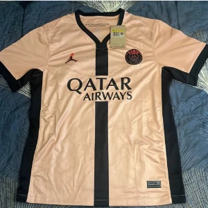 PSG fotbollströja rosa/svart Jordan - Snygg PSG fotbollströja i ljusrosa med svarta detaljer och vertikal rand framtill. Tröjan har korta ärmar, svart V-ringad krage och Paris Saint-Germain-logga på bröstet. Tillverkad i lätt och ventilerande polyester, perfekt för match eller träning.