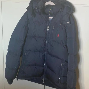 Mörkblå dunjacka från Polo Ralph Lauren - Säljer en mörkblå Ralph Lauren dun jacka som är helt perfekt till vintern. Jackan användes endast under förra vinter och ett fåtal defekter förekommer. Kvitto och äkthetsbevis finns, nypris 4899 kr. Priset är ej hugget i sten och vid en snabb affär kan ett bra pris tilldelas.     Vid frågor eller funderingar är det bara att skriva