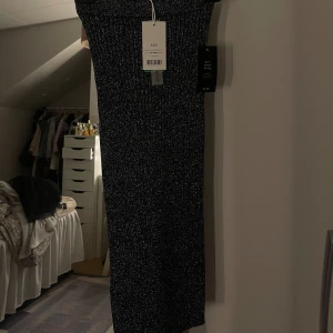 Glittrig svart/grå långkjol från NA-KD - Svart/grå glittrig kjol i ribbat stretchtyg, storlek XXS( passar xs). Kjolen är helt ny med lappen kvar och säljs endast på grund av fel storlek. Den är figurnära och bekväm med stretchigt material som formar sig fint efter kroppen.