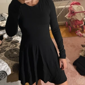 Svart långärmad kortklänning - Enkel och stilren svart klänning med lång ärm och rund hals. Modellen är figurnära upptill och har en lätt utställd kjol som ger en snygg siluett. Perfekt för dig som gillar minimalistisk stil och vill ha något som funkar till många tillfällen.