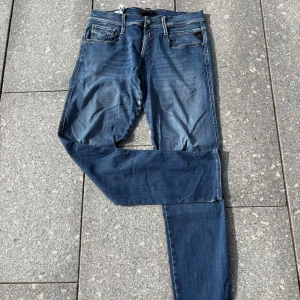 Blå Replay Anbass jeans herr - Säljer ett par klassiska blå Replay Anbass jeans med fem fickor och snygga slitningar. Jeansen har normal passform och raka ben, med detaljerade sömmar och logga på bakfickan. Perfekta för dig som gillar en tidlös denimlook.