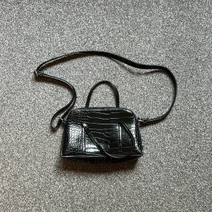 Svart axelväska med krokoprägling - Snygg svart axelväska med krokodilpräglad yta och både axelrem och handtag. Väskan har dragkedja upptill och är perfekt för att bära det viktigaste. Stilren och trendig modell som passar till många outfits.