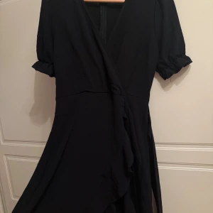 Svart omlottklänning med knyt - Svart omlottklänning med korta puffärmar och volangdetaljer. Klänningen har v-ringad hals och knyt i midjan för en snygg siluett. 