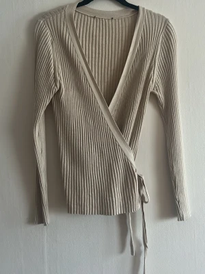 Beige ribbad omlottkofta - Säljer en beige ribbad omlottkofta med långa ärmar och knytband i midjan. Koftan har djup v-ringning och är tillverkad i ett mjukt, stretchigt material som sitter snyggt på kroppen. Perfekt för lager-på-lager och enkel att styla till olika outfits.