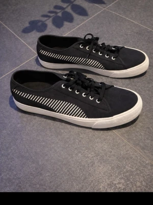 Sneakers skor  - Säljer ett par svarta Puma sneakers med vita snedställda ränder på sidorna. Skorna har snörning, vit platt sula och rund tå. Ovandelen är i textil och sulan är i gummi. Perfekta för dig som gillar stilrena och sportiga skor.sneakers #skor #fashion #stilren