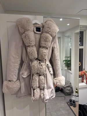 Beige parkas med fuskpäls från Chelsea  - Säljer en beige parkas från Chelsea med fluffig fuskpäls runt huva, krage och är soppar. Jackan har snörning i midjan, stora fickor och stängs med både dragkedja och knappar. Perfekt för kalla dagar och riktigt mysig att bära. Pris kan diskuteras 