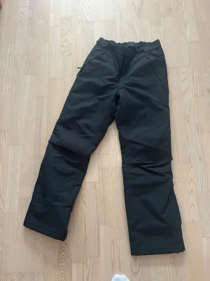 Svarta täckbyxor från North Bend - Säljer ett par svarta täckbyxor från North Bend med elastisk midja och justerbart kardborreband. Byxorna är vattentäta med tejpade sömmar och har dragkedjor vid fickorna. Hela och rena.