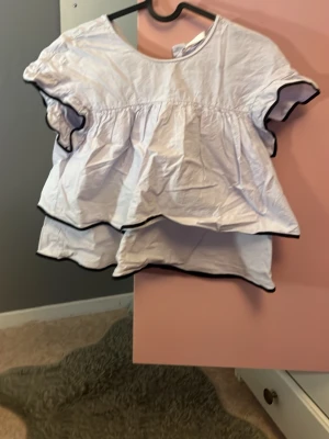 Vit volangblus med svarta kanter - Supersöt vit blus med dubbla volanger och svarta vågiga kanter. Blusen har korta ärmar och en luftig, loose passform som ger en avslappnad vibe. Perfekt till jeans eller kjol för en trendig look.