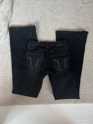 Svarta bootcut jeans med detaljer - Säljer ett par svarta bootcut jeans med snygga sömmar och dekorativa nitar på bakfickorna. Jeansen har klassisk femficksmodell, normal passform och är tillverkade i mjukt jeansmaterial. Perfekta för dig som gillar en tidlös look med lite extra edge.