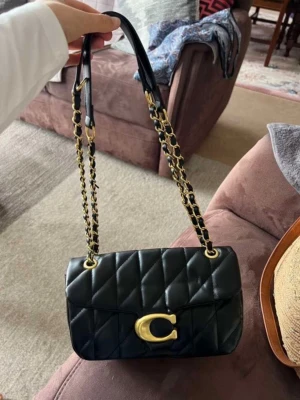 Svart quiltad axelväska från Coach - Snygg svart axelväska från Coach i quiltat skinn med guldfärgade kedjedetaljer och stort C-spänne framtill. Väskan har flera fack inuti och en lyxig känsla med både läder och metall i axelremmen. Perfekt för dig som vill ha en trendig och stilren väska med exklusiva detaljer.