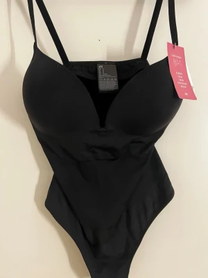 Svart shaping-body från H&M - Svart shaping-body från H&M med tunna axelband och vadderade kupor. Snygg och stilren modell som ger en smickrande siluett. Tillverkad i stretchigt syntetmaterial för en bekväm passform. Den är helt ny med prislapp 