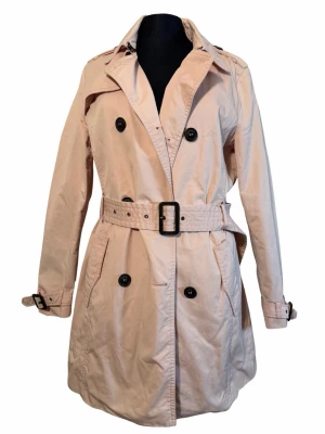 Ljusrosa trenchcoat med bälte ESPRIT - Snygg ljusbeige trenchcoat med dubbelknäppning och bälte i midjan. Jackan har stora svarta knappar, klassisk krage och justerbara spännen vid ärmsluten. Perfekt för dig som vill ha en stilren och tidlös look. Mycket bra skick från ESPRIT.