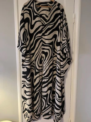 Zebramönstrad långklänning med knappar - Säljer en flowy långklänning med svartvitt zebramönster och v-ringning. Klänningen har vida ärmar och knappar framtill, vilket ger en chill och unik look. Materialet känns lätt och svalt, perfekt för dig som vill sticka ut. Skicket är perfekt och passar uppknäppt över svart t- shirt och tajts och lika bra över bikinin. 