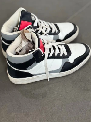 Svarta och vita high top sneakers - Säljer ett par high top sneakers i svart, vitt och grått med coola detaljer och en röd patch på plösen. Skorna har snörning, rund tå och platt sula. Materialet är syntet och textil, perfekta för dig som gillar streetstyle.