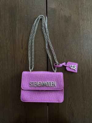 Rosa axelväska från Steve Madden - Rosa  axelväska från Steve Madden med präglat logomönster över hela väskan. Väskan har en lång silverfärgad kedja somrar att ha lång och kort. 