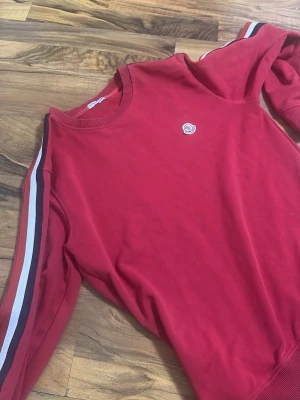 Moncler tröja kofta - Röd sweatshirt från moncler med rund hals och ribbade muddar. Ärmarna har snygga ränder i vitt, svart och rött längs sidorna. Liten broderad logga på bröstet. Perfekt för dig som gillar sportig och avslappnad stil.