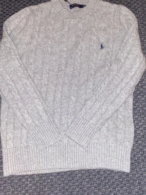 Grå kabelstickad tröja Polo Ralph Lauren - Snygg grå kabelstickad tröja från Polo Ralph Lauren med rund hals och diskret blå broderad logga på bröstet. Tröjan har långärmade ribbade muddar och är gjord i mjukt stickat material som passar perfekt för kyliga dagar.