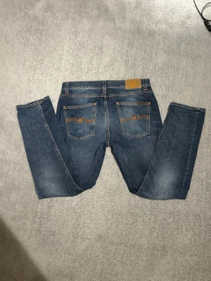Nudie Jeans Lean Dean 31/30 - Säljer ett par klassiska blå jeans från Nudie Jeans med snygga orangea sömmar och broderad logga på bakfickan. Modellen är Lean Dean dvs slim fit. Jeansen är tillverkade i slitstarkt bomullsmaterial och har fem fickor samt knappgylf.
