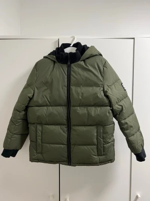 Grön pufferjacka från Diesel  - Snygg grön pufferjacka från Diesel med huva och svart dragkedja. Jackan har stora fickor framtill, ribbade muddar och tydligt Diesel-tryck i gult på ryggen. Materialet är 100% nylon och jackan har en loose passform som ger en chill vibe.