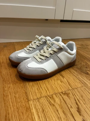 Vita och grå sneakers med mockadetaljer - Säljer ett par stilrena sneakers i vitt och ljusgrått med beige snörning och bruna gummisulor. Skorna har paneler i mocka och släta partier i syntet, samt klassisk rund tå och platt sula. Perfekta för dig som gillar minimalistisk och clean stil.