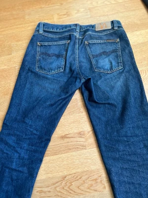 Blå jeans från Nudie Jeans Co gritty jacksson - Säljer ett par klassiska blå jeans från Nudie Jeans Co med fem fickor, snygga kontrastsömmar och logga på bakfickan. Jeansen har rak passform och är tillverkade i slitstarkt bomullsmaterial. Perfekt för dig som gillar tidlös denimstil.