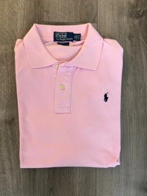 Rosa pikétröja från Polo Ralph Lauren - Snygg rosa pikétröja från Polo Ralph Lauren i storlek XXL. Klassisk krage, två knappar och den ikoniska mörkblå broderade loggan på bröstet. Tillverkad i 100% bomull och har en relaxed custom fit. Storleken är XXL men eftersom den har tvättats ett fåtal gånger så har den krympt, så den passar för dig som har M