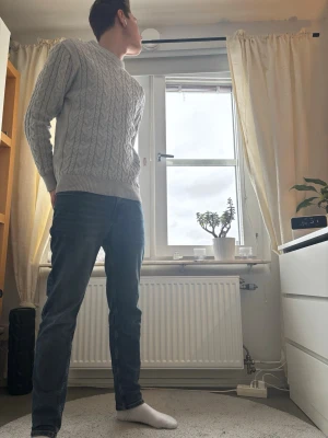 Ljusgrå kabelstickad tröja - Säljer en ljusgrå kabelstickad tröja med rund hals och ribbade muddar. Tröjan har ett klassiskt flätmönster och är långärmad, perfekt för kyligare dagar. Passformen är normal och materialet känns mjukt och bekvämt.