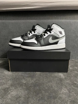 Nike Air Jordan 1 Mid svart/vit - Säljer ett par Nike Air Jordan 1 Mid i svart och vitt skinn med grå swoosh och klassisk Jordan-logga på sidan. Skorna har snörning, rund tå och en platt sula. Perfekta för dig som gillar streetstyle och vill ha en ikonisk sneaker i garderoben. Helt nya inte ens använda köpte fel storlek därför säljer jag dom 