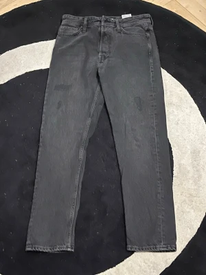 Jack and Jone svarta jeans 28/30 - Säljer ett par svarta jeans från jack and jones i storlek 28/30. Jeansen har en loose passform med raka ben och klassisk femficksdesign. Materialet är slitstarkt jeans och färgen är en snygg mörkgråsvart ton. Perfekta för dig som gillar avslappnad stil. Passform loose chris.
