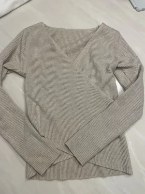 Beige ribbad omlottopp med lång ärm - Säljer en beige ribbad omlottopp med lång ärm och v-ringad hals.  Liten fläck, kan tvätta bort innan jag skickar paketet. 