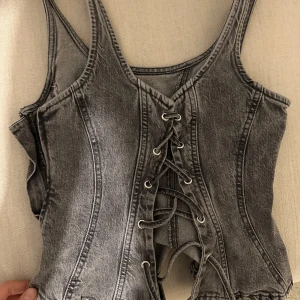 Grå denimkorsett med snörning från Gina tricot - Säljer en grå korsettopp i denim-look med snygg snörning framtill. Toppen har breda axelband och markerade sömmar som ger en cool och edgy vibe. Perfekt att styla med jeans eller kjol för en trendig outfit.