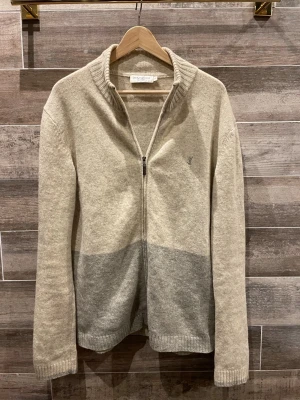 Yves Saint Laurent tröja  - Säljer en beige och ljusgrå kofta från Yves Saint Laurent. Koftan har hel dragkedja framtill, ribbad krage och muddar samt diskret YSL-logga broderad på bröstet. Tillverkad i mjuk ullblandning, perfekt för kyliga dagar. Nypris 6000kr! 