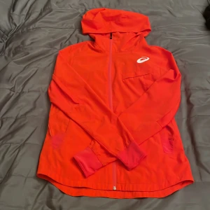 Röd vindjacka från Asics med huva - Säljer en snygg röd vindjacka från Asics i storlek M. Jackan har huva, hel dragkedja framtill och långa ärmar med ribbade muddar. Materialet är lätt och syntetiskt, perfekt för aktiva dagar. Asics-logga i vitt på bröstet ger en sportig vibe.