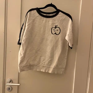 Vit kortärmad tröja Bobo Choses - Vit kortärmad tröja från Bobo Choses med svarta detaljer vid hals och ärmar. Framsidan har ett tryckt äpple med ett sött ansikte. Tröjan har en avslappnad passform och är gjord i mjuk bomull, perfekt för chill dagar.